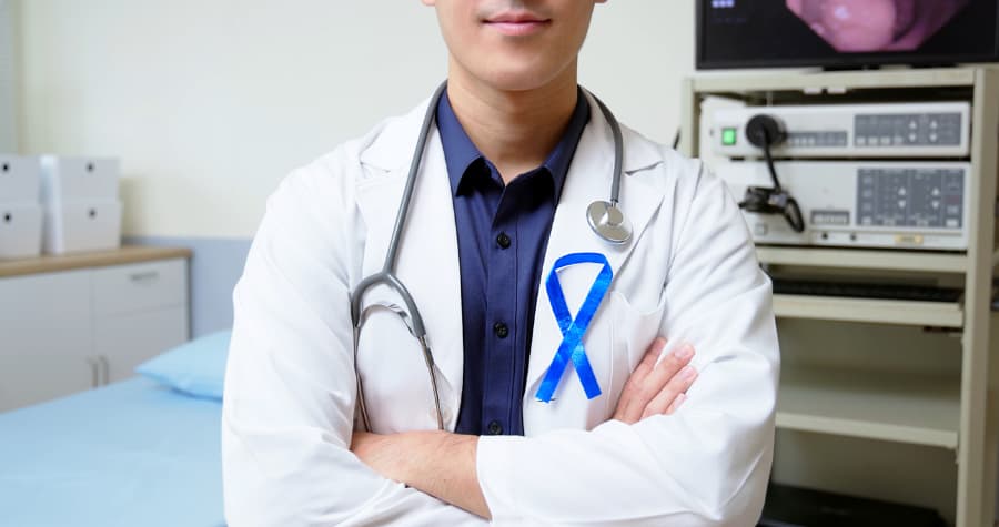 Physician with arms folded wearing white medical coat with blue ribbon